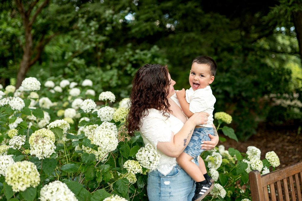 A mother lifts her smiling toddler among white hydrangea blooms in a lush green garden setting.