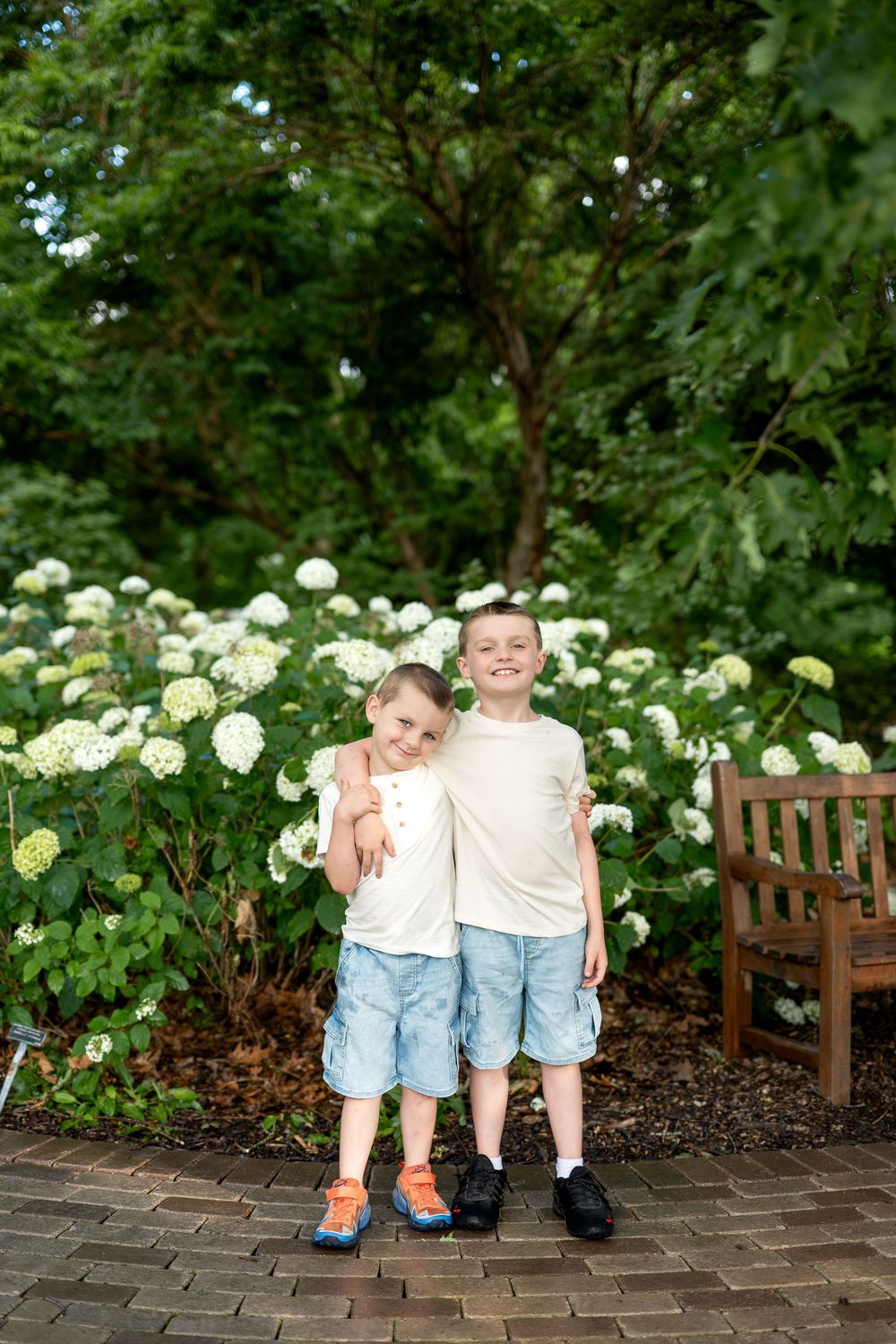 Two young boys in matching white shirts and blue shorts pose together in front of white hydrangeas in a lush garden.