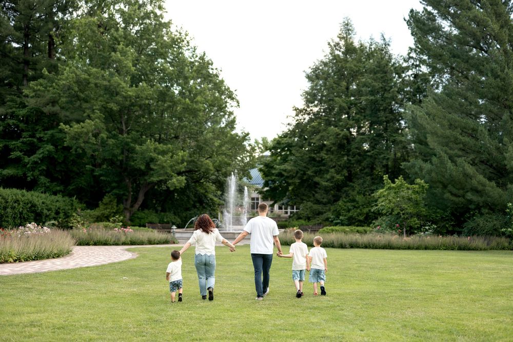 Family of four holding hands walking through a lush green park toward a fountain on a summer day.