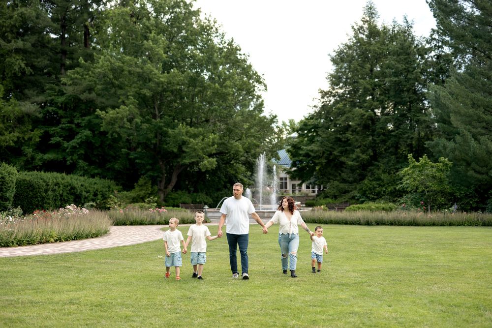 Family of five holding hands walking on green lawn with fountain and trees in background.