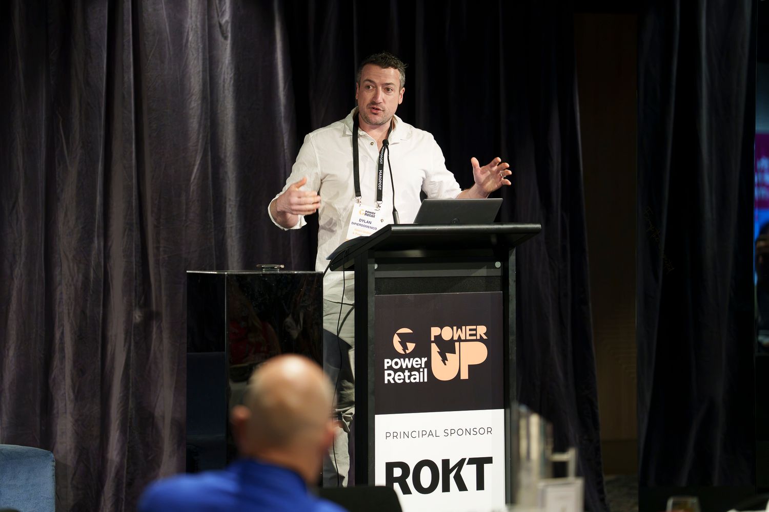 Speaker presenting at Power Retail Power Up conference with ROKT principal sponsor podium in modern event venue.