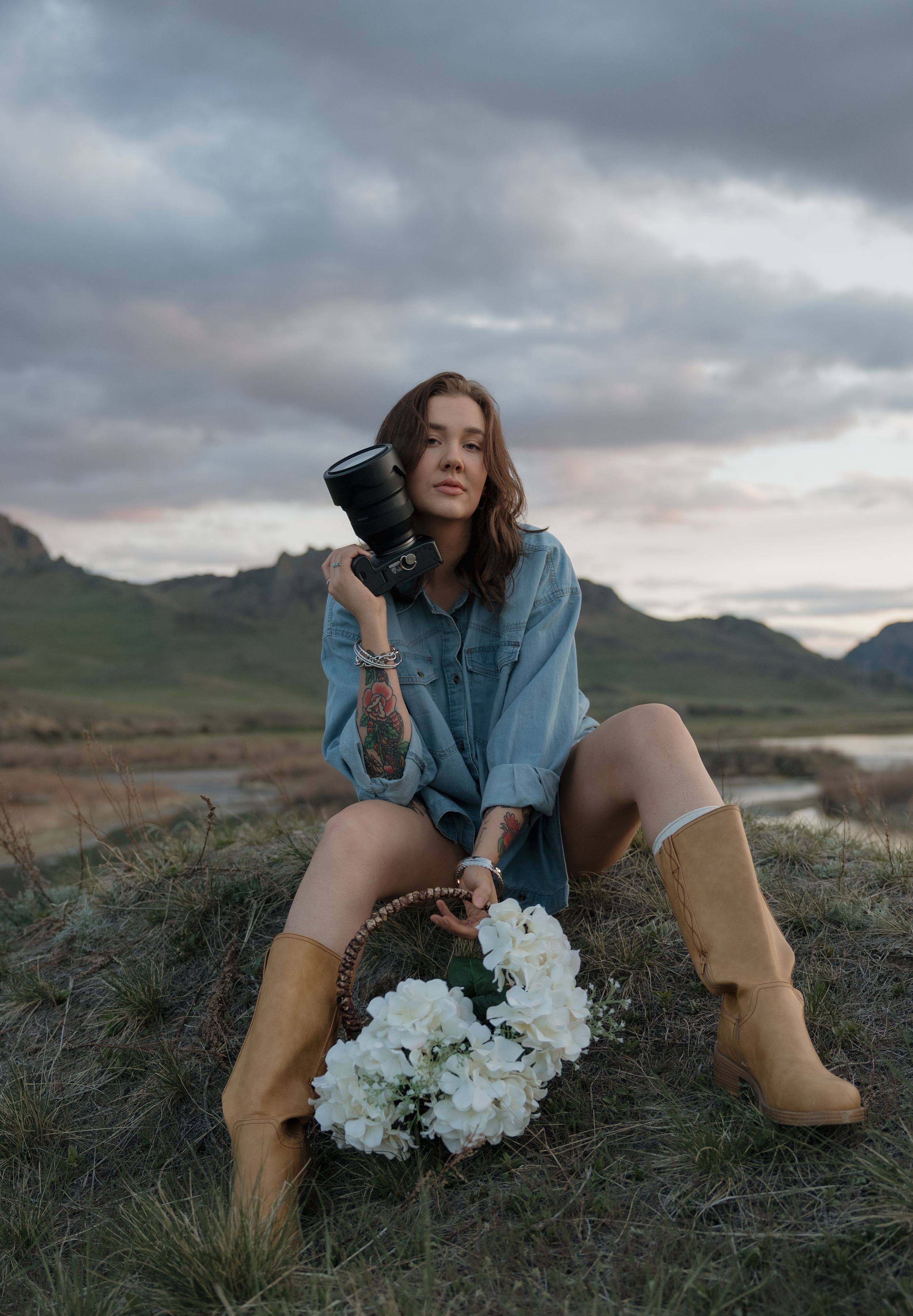 Young woman in denim shirt and tan boots sits in field holding camera with white flowers.