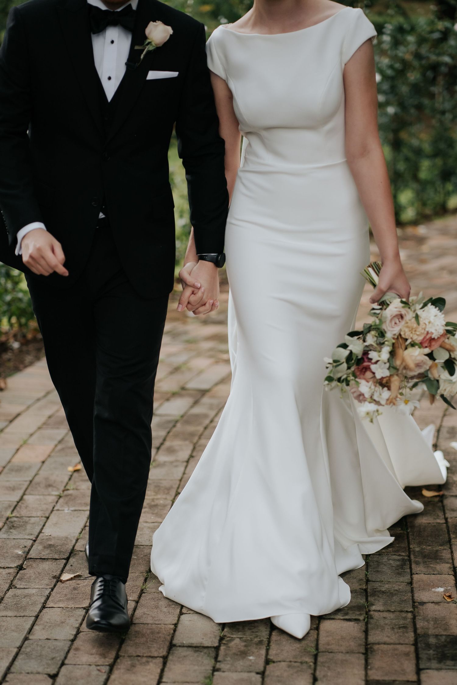 Bride in white mermaid gown holds bouquet while walking with groom in black tuxedo on brick path.