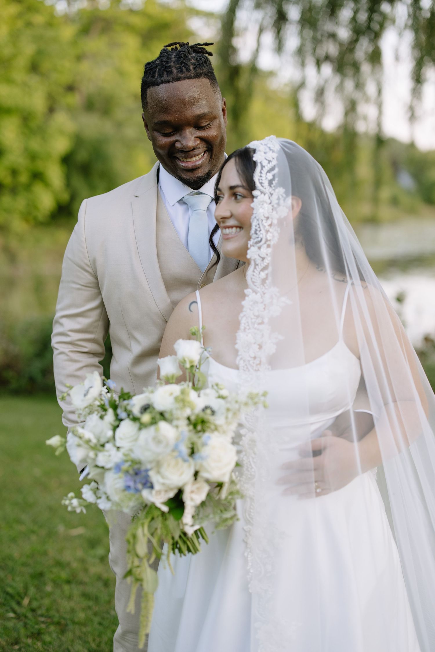 Smiling interracial newlywed couple outdoors, groom in beige suit, bride in white gown with veil holding blue floral bouquet.