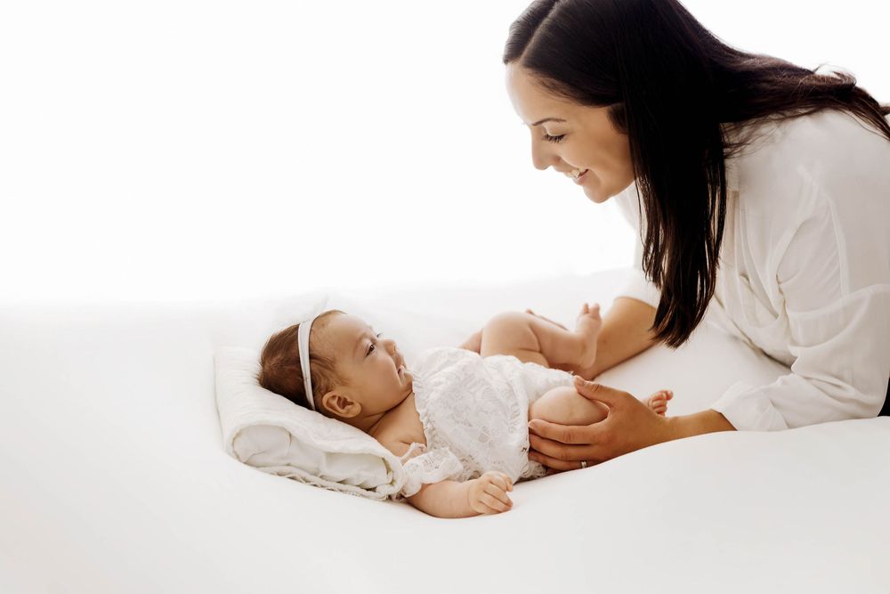 A mother smiles at her newborn baby lying on a white bed in matching white outfits.