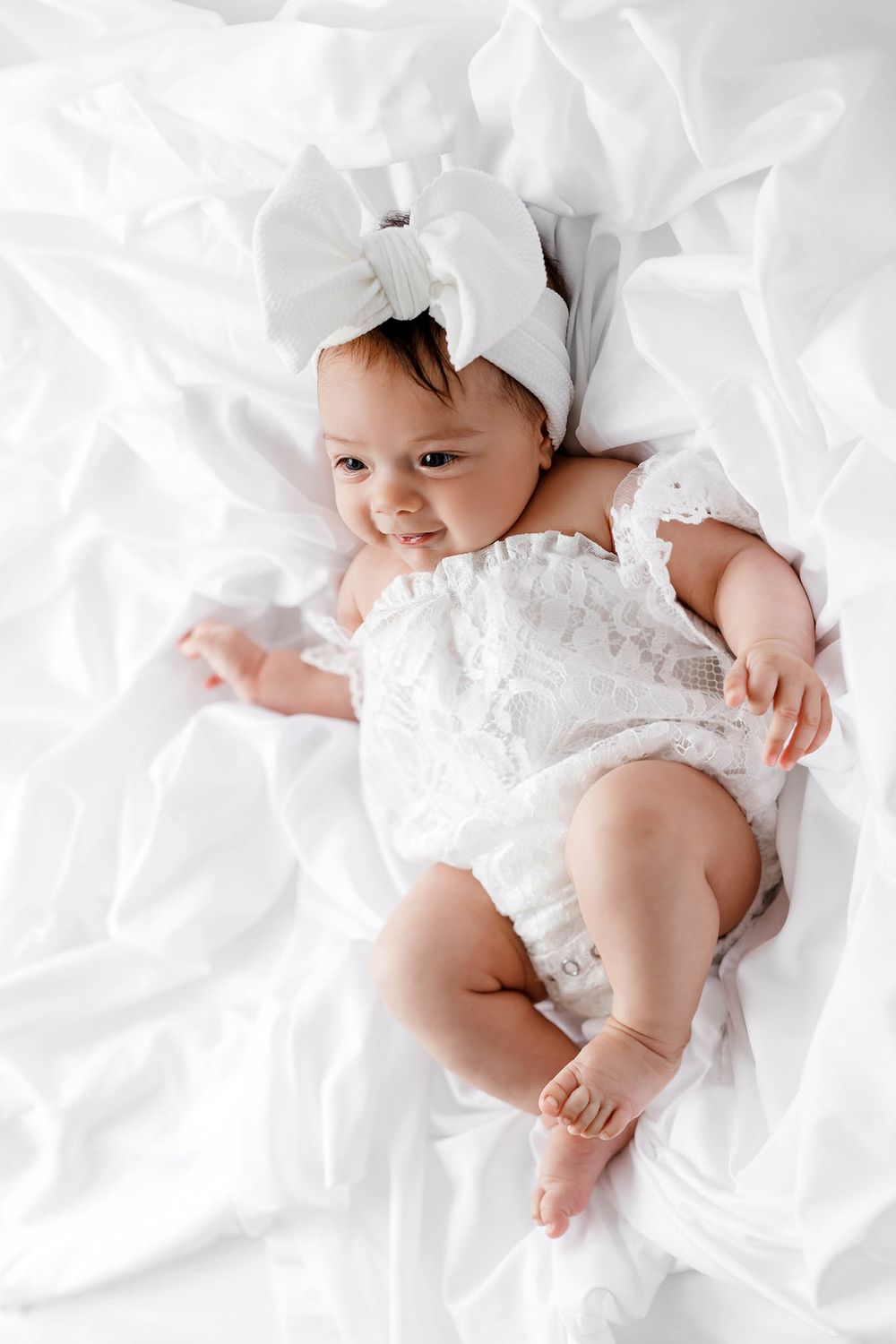 Smiling baby girl in white lace romper and bow headband lying on white bedding.