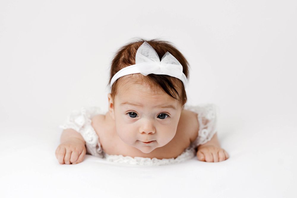 Adorable baby girl in white lace dress and bow headband doing tummy time on white background.