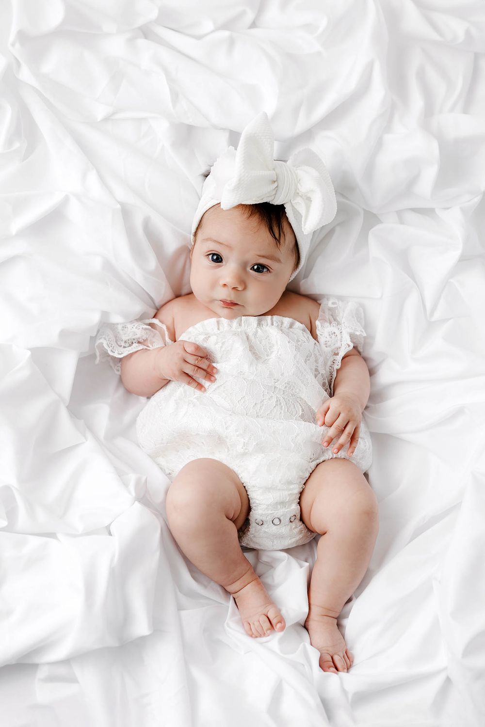 Adorable baby girl in white lace romper and bow headband lying on white bedding, looking at camera.