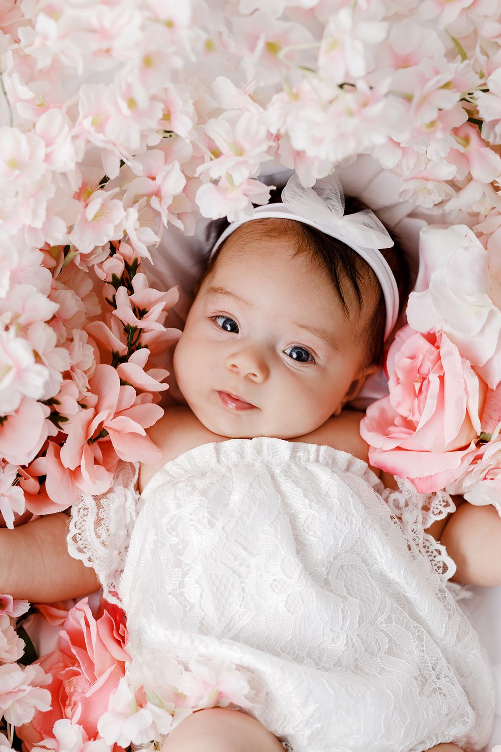 Adorable baby girl in white lace dress and bow headband lying among pink roses and blossoms.