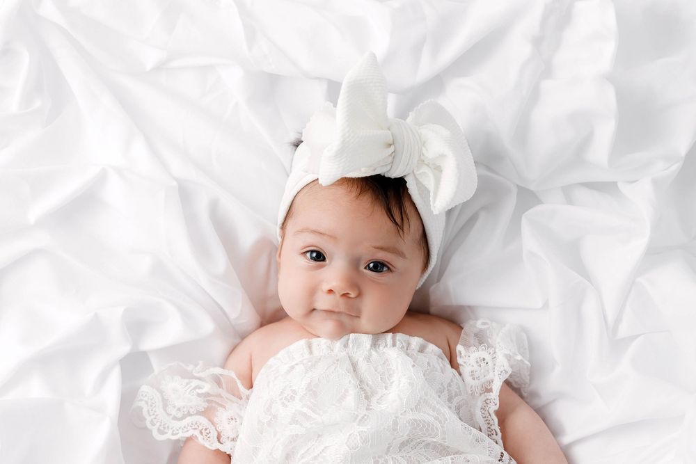 Adorable baby girl in white lace outfit and bow headband lying on white sheets.