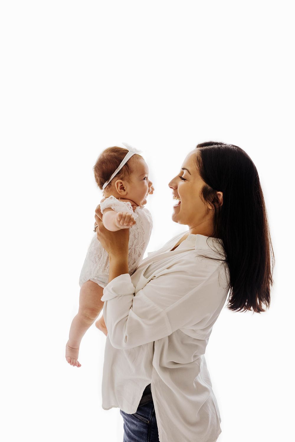 Happy mother lifting and smiling at her baby girl in white outfit against bright white background.
