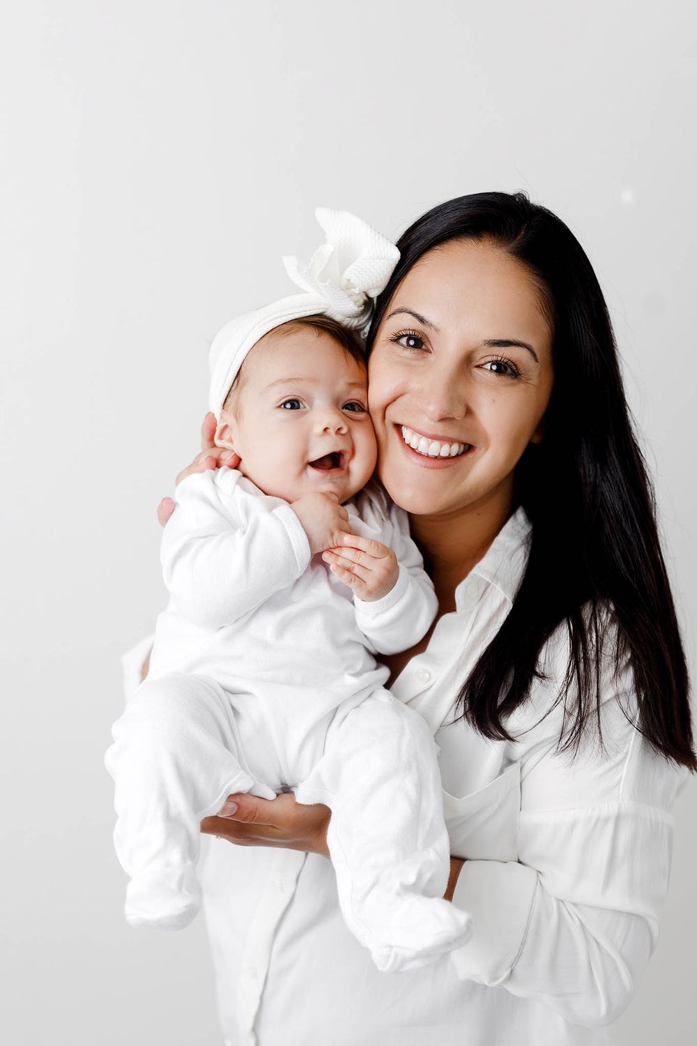 Smiling mother in white shirt holds her happy baby girl wearing a white bow headband.