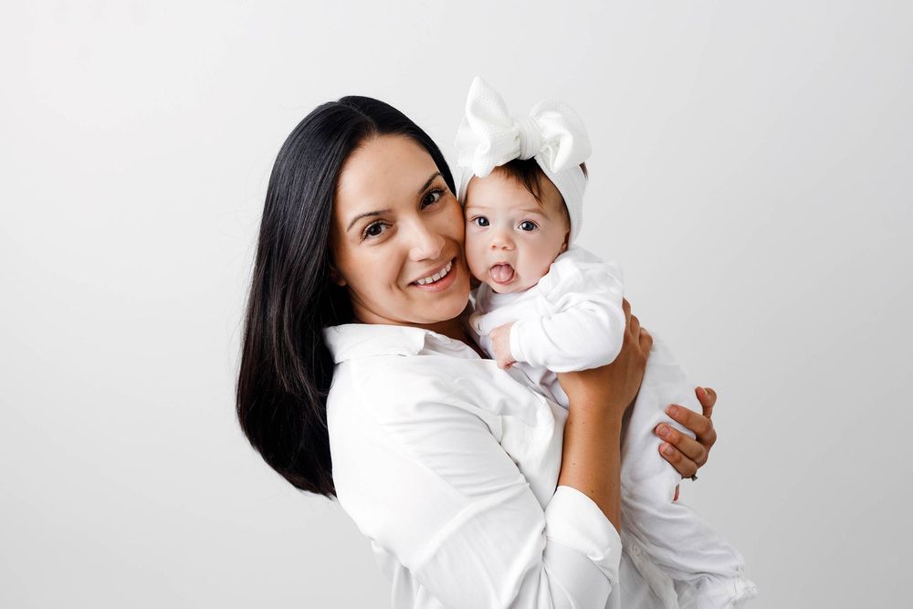 Smiling dark-haired woman in white shirt holding adorable baby girl wearing white bow headband against light background.