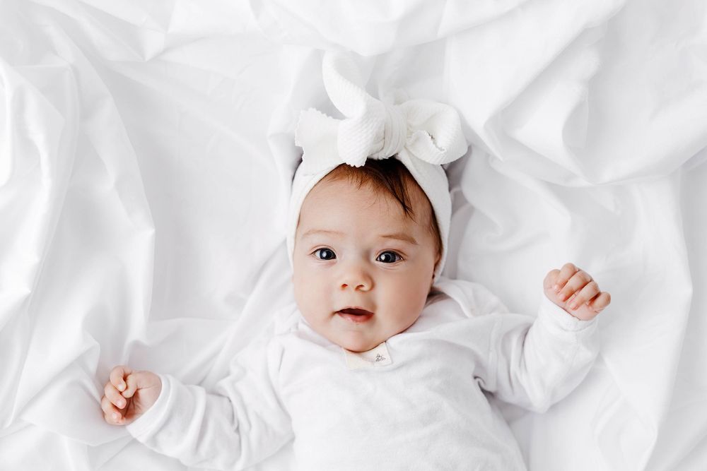 Adorable baby in white outfit and bow headband lying on white bedding looking at camera.