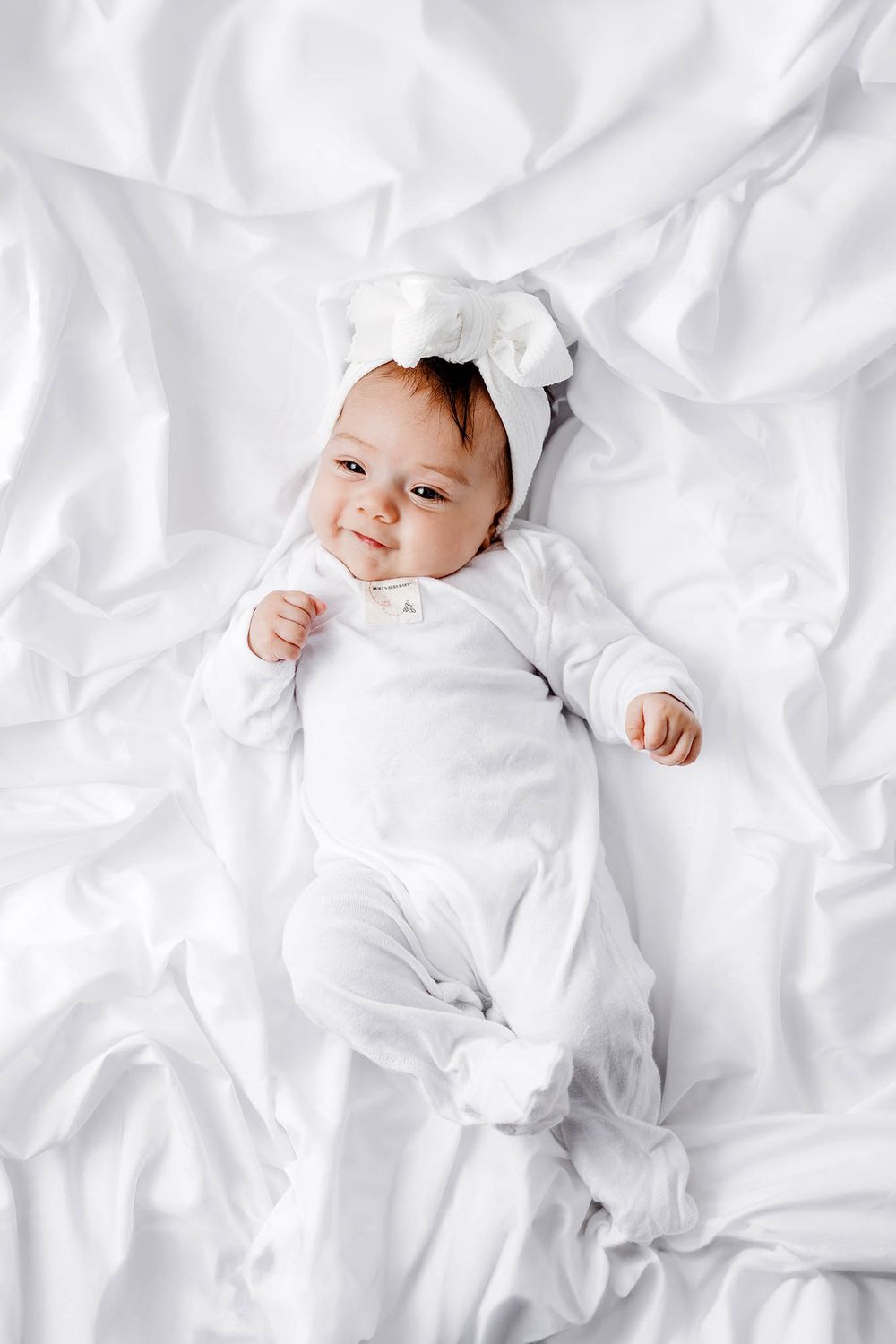 Adorable baby in white onesie and bow headband lying on white sheets smiling at camera.