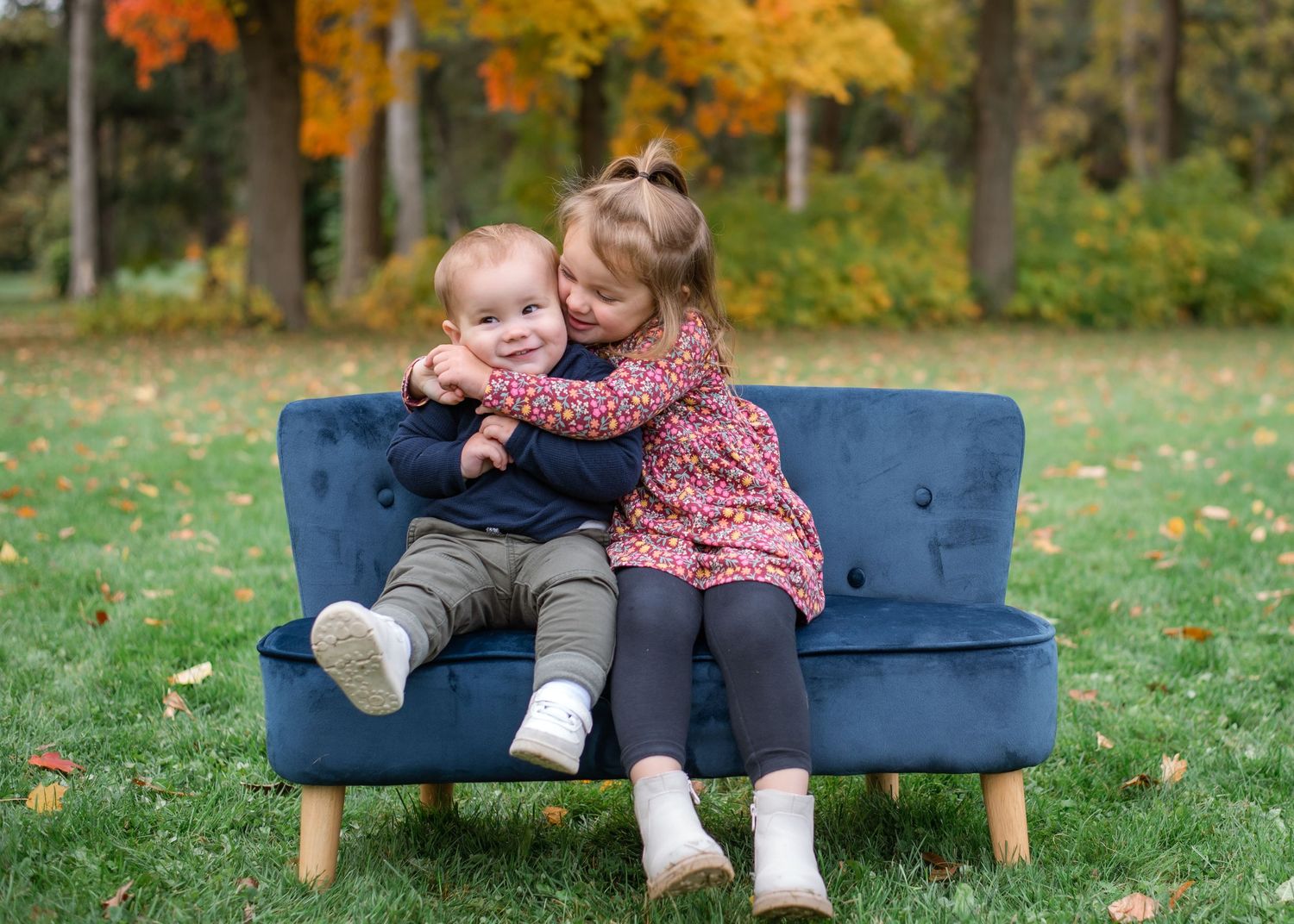 Two young children sitting on a small blue sofa outdoors, with a girl hugging a baby boy among autumn foliage.