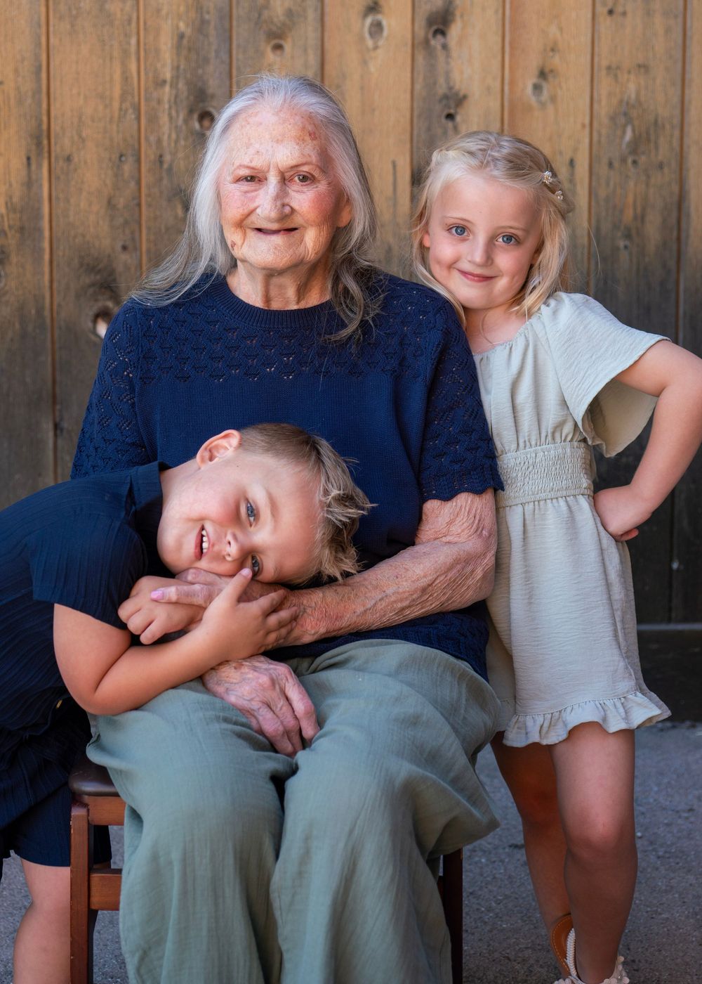 Elderly woman smiling warmly with two young grandchildren, boy and girl, posing together in front of a wooden wall.