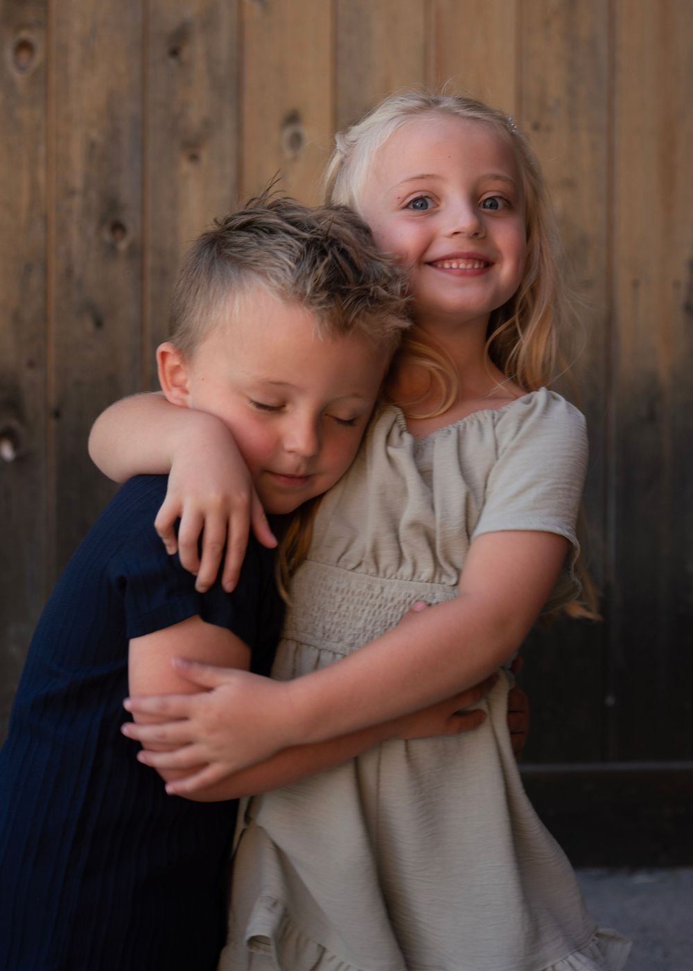 Two young children sharing a warm hug, a smiling blonde girl in a beige dress embracing a boy in a navy shirt outdoors.