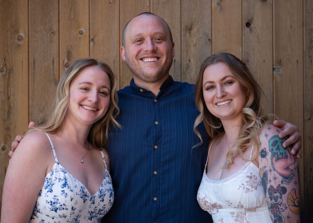 Three smiling adults posing together in front of a rustic wooden wall at a casual outdoor gathering.