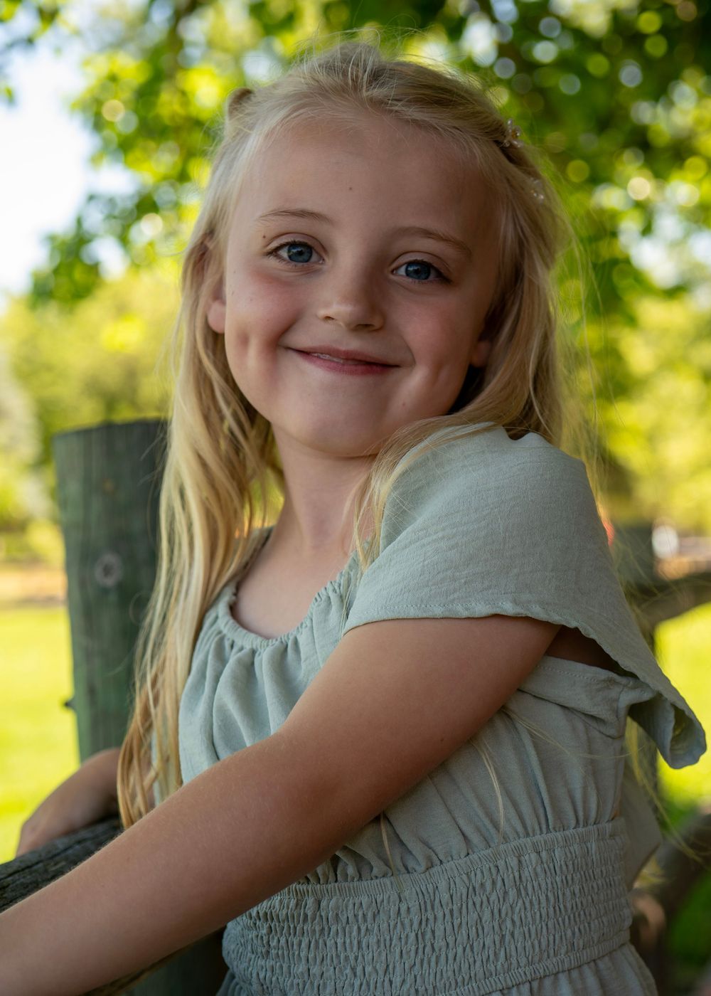 Young blonde girl smiling outdoors in sage green dress, leaning on wooden fence with green trees in background.