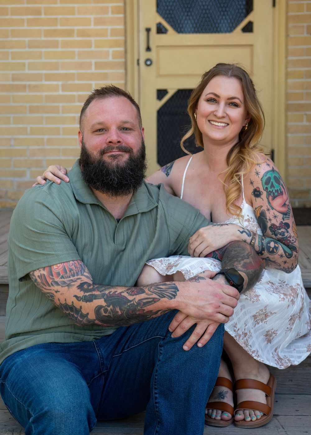 Tattooed couple sitting together on steps in front of a brick building, smiling warmly at the camera.