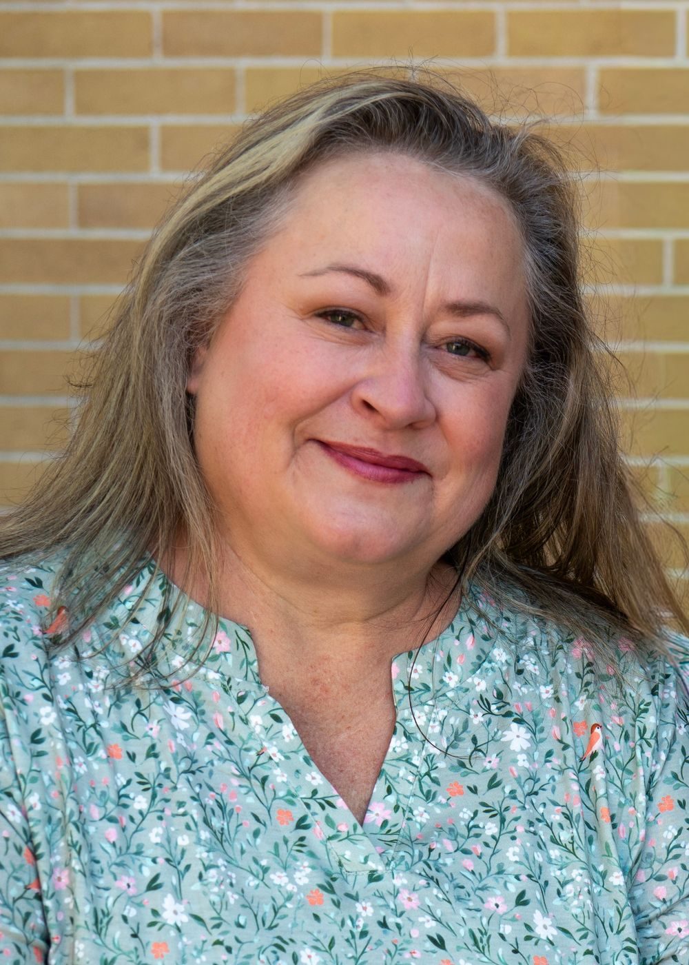 A smiling middle-aged woman with blonde hair wearing a floral blouse poses in front of a brick wall.