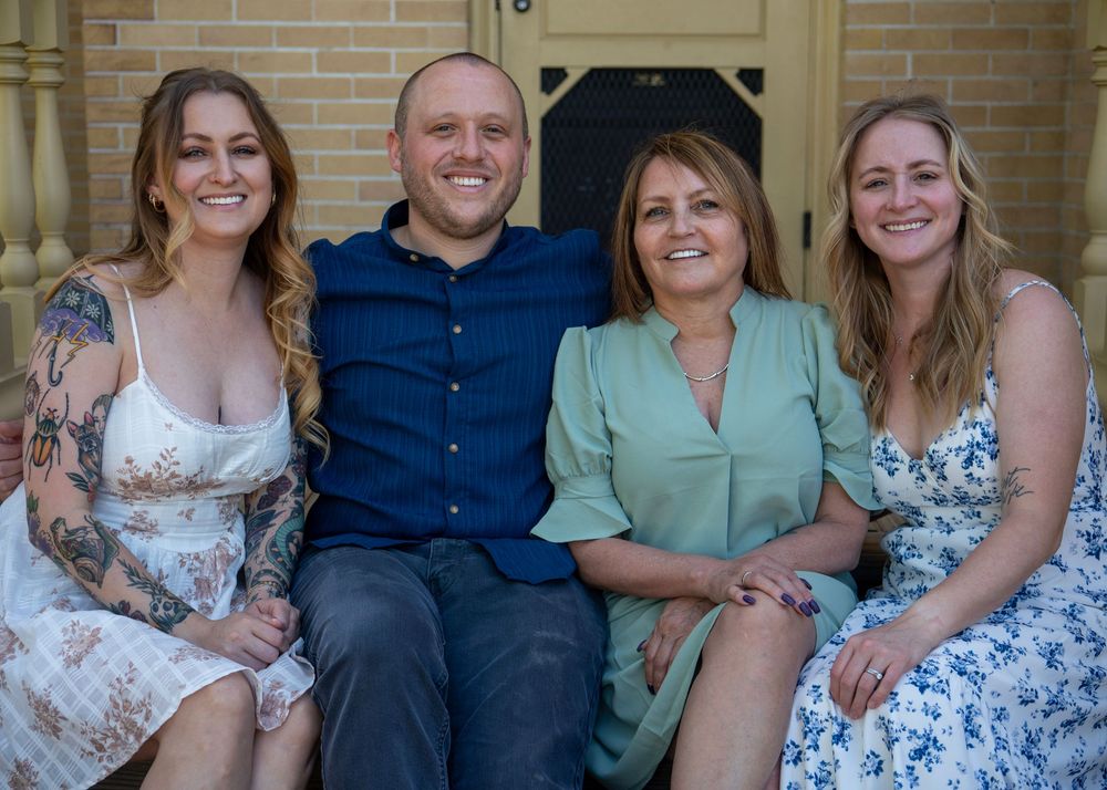A woman with three adult children sitting and smiling together for a family portrait on outdoor steps.