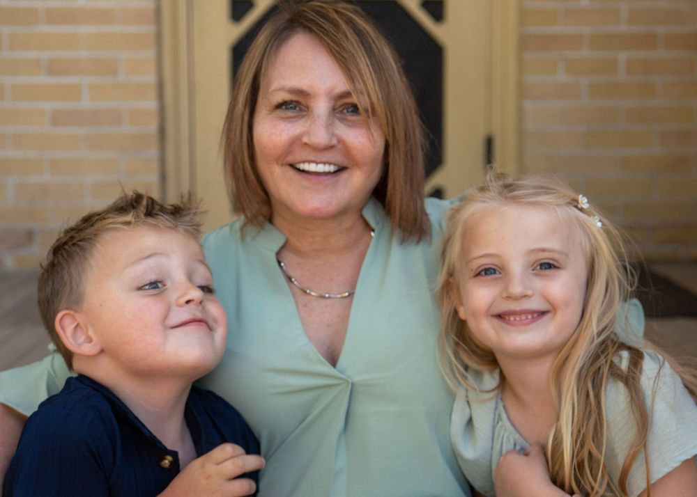 A smiling woman with short brown hair poses with two young blonde children in front of a brick building.