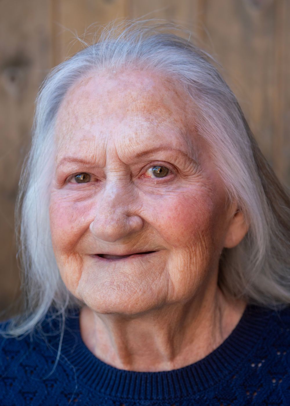 Elderly woman with long gray hair and a warm smile wearing a navy blue sweater against a wooden background.