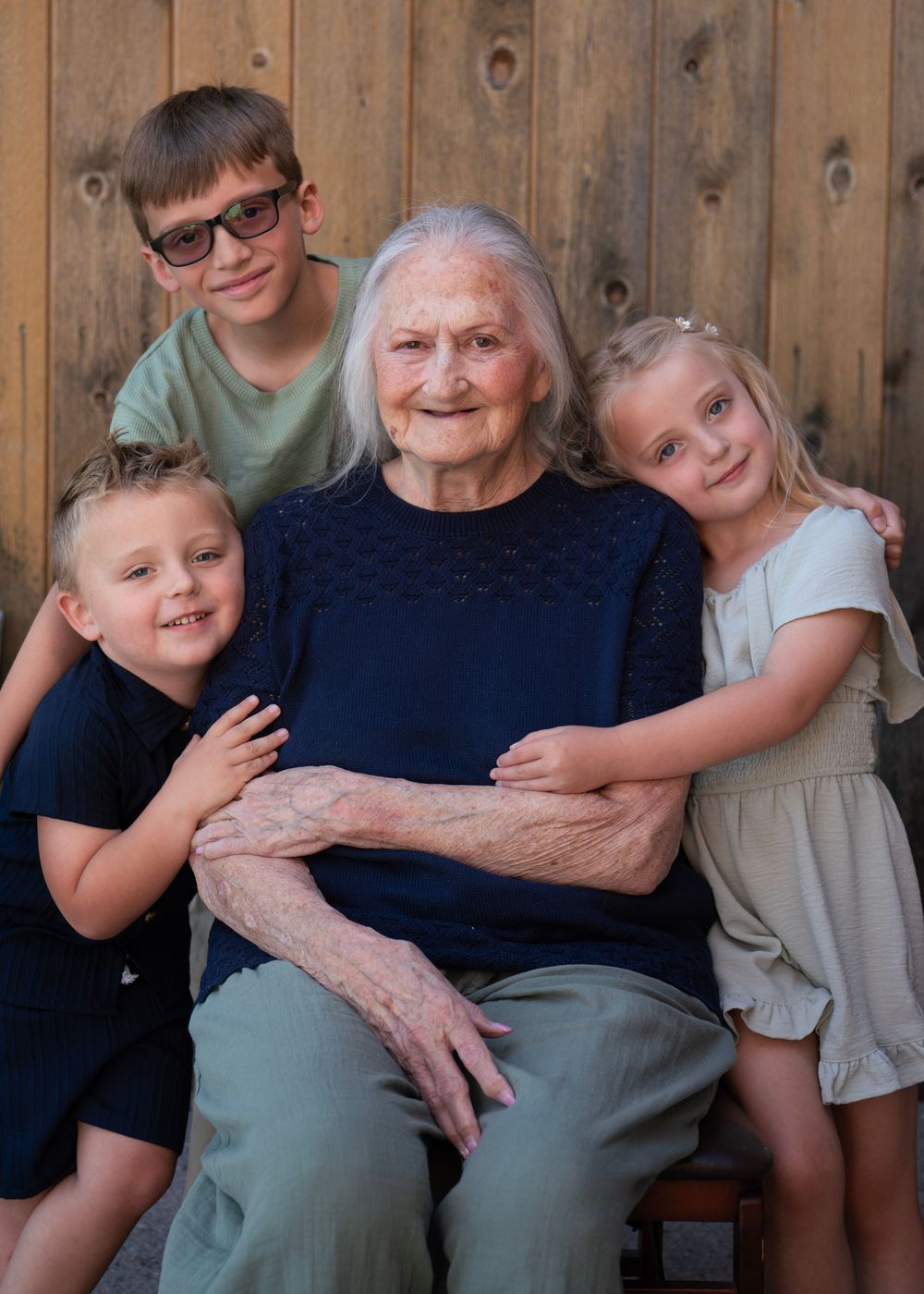 Elderly woman seated with three young grandchildren hugging her, posing for a family portrait against a wooden wall.