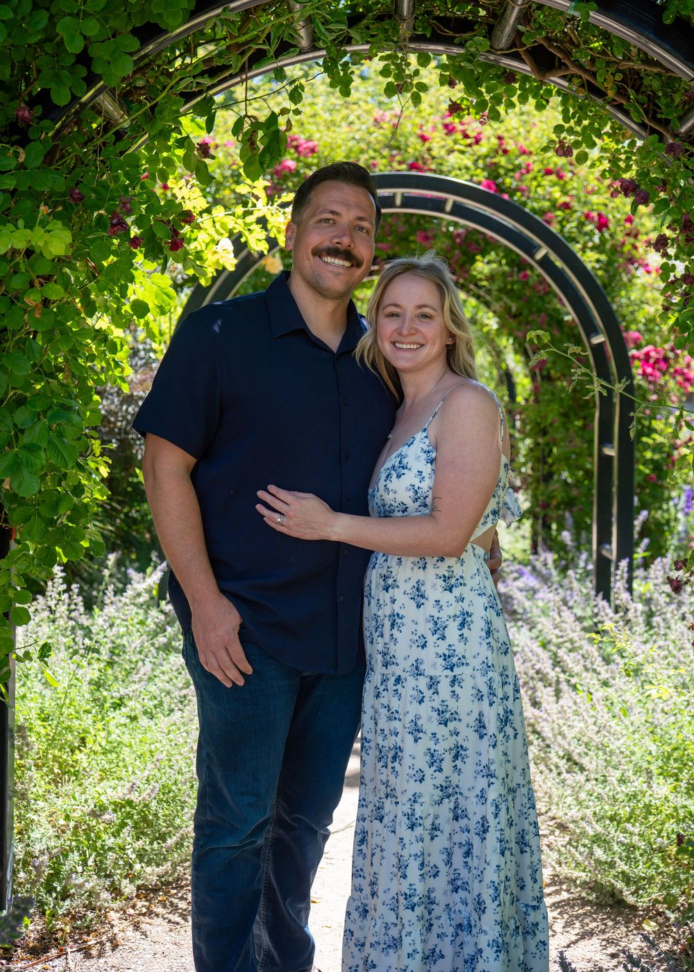 A smiling couple posing under a lush floral garden archway surrounded by colorful blooming flowers.