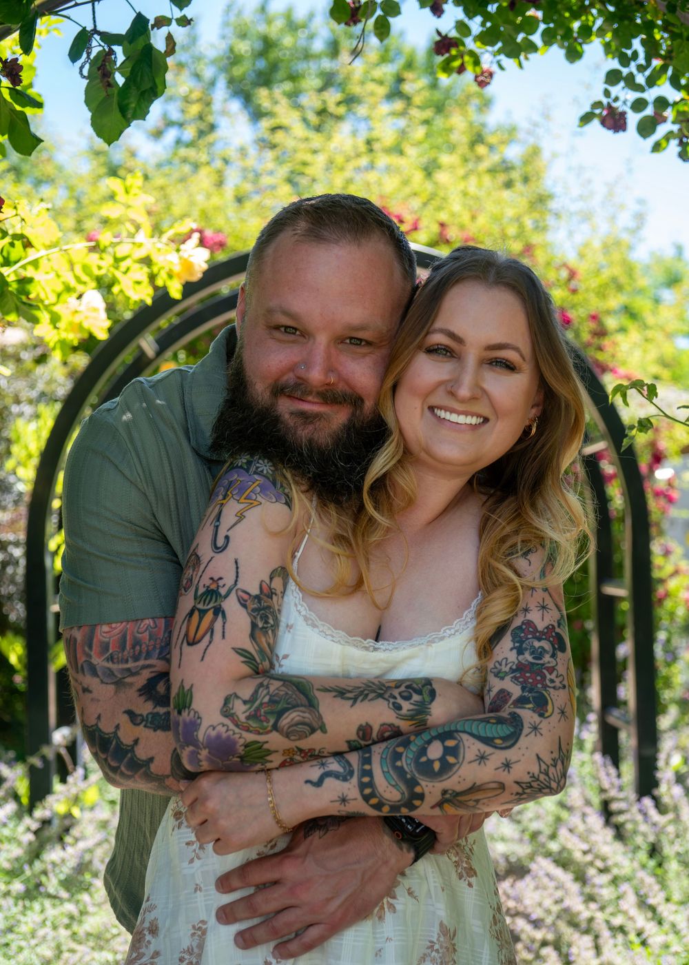A tattooed couple embraces and smiles in a lush garden with a floral arch, the woman in a white dress and man in green shirt.