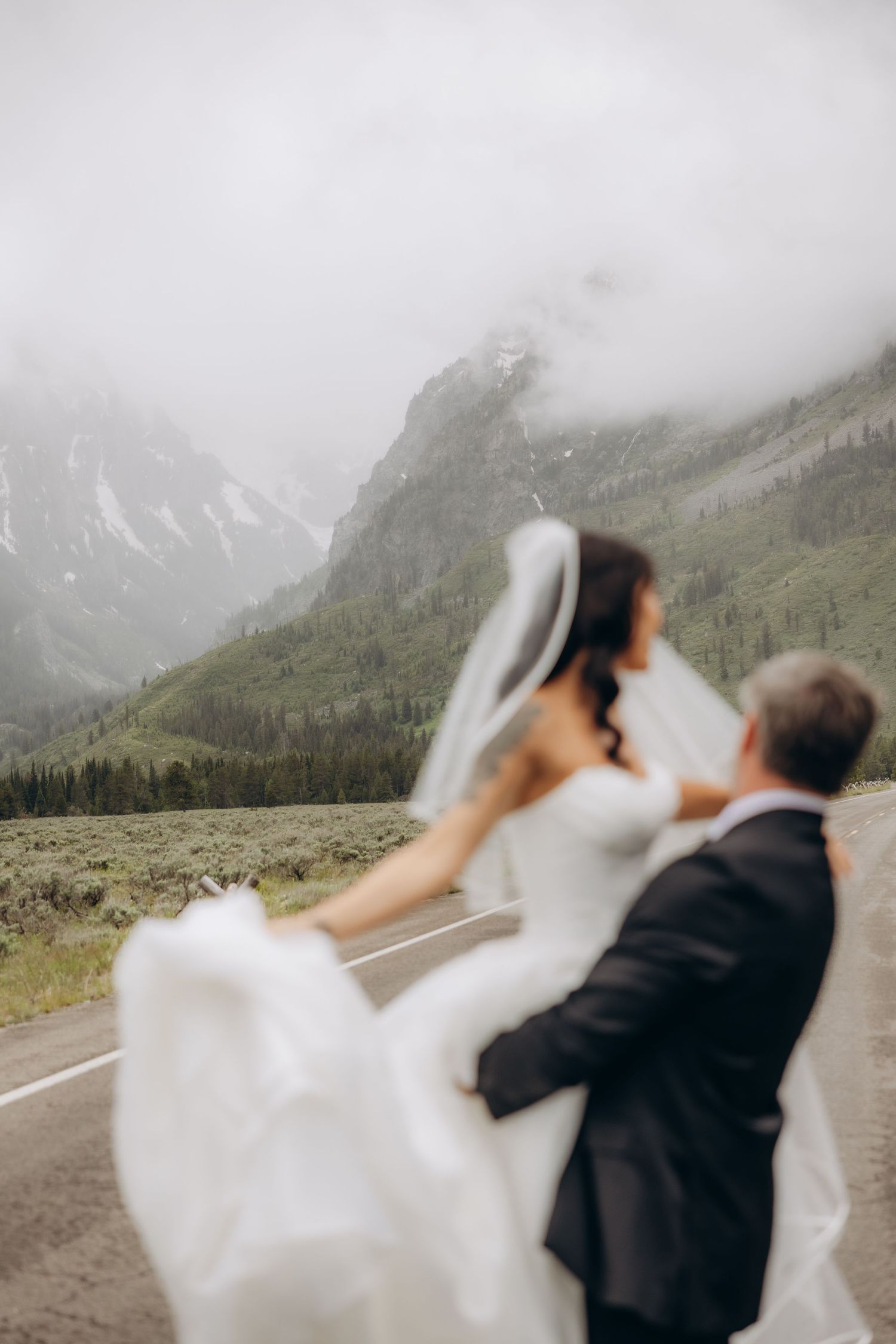 Bride and groom share a romantic moment on a mountain road, with misty peaks and green forests in the background.