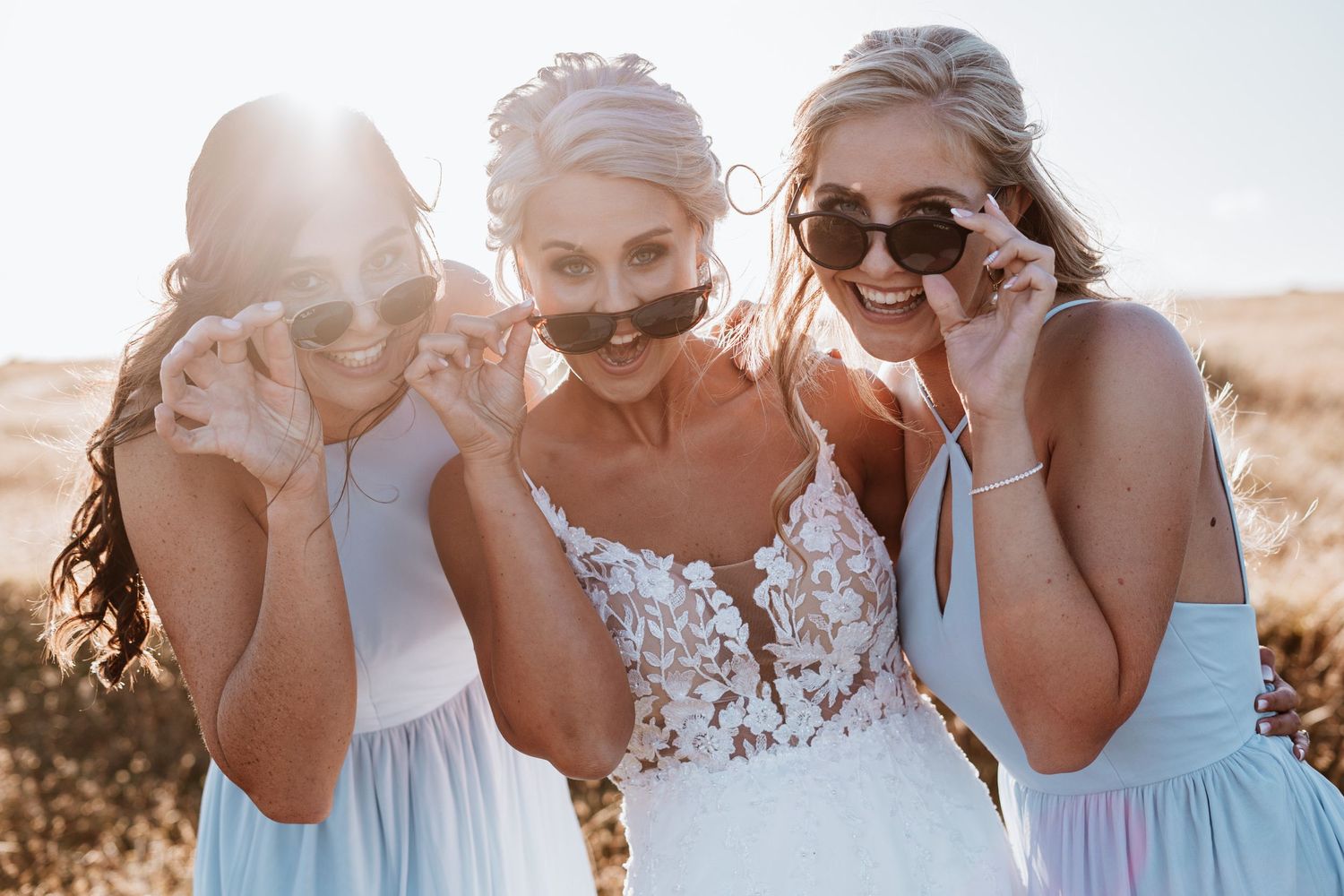 Bride and two bridesmaids in light blue dresses posing with sunglasses in sunny outdoor field.