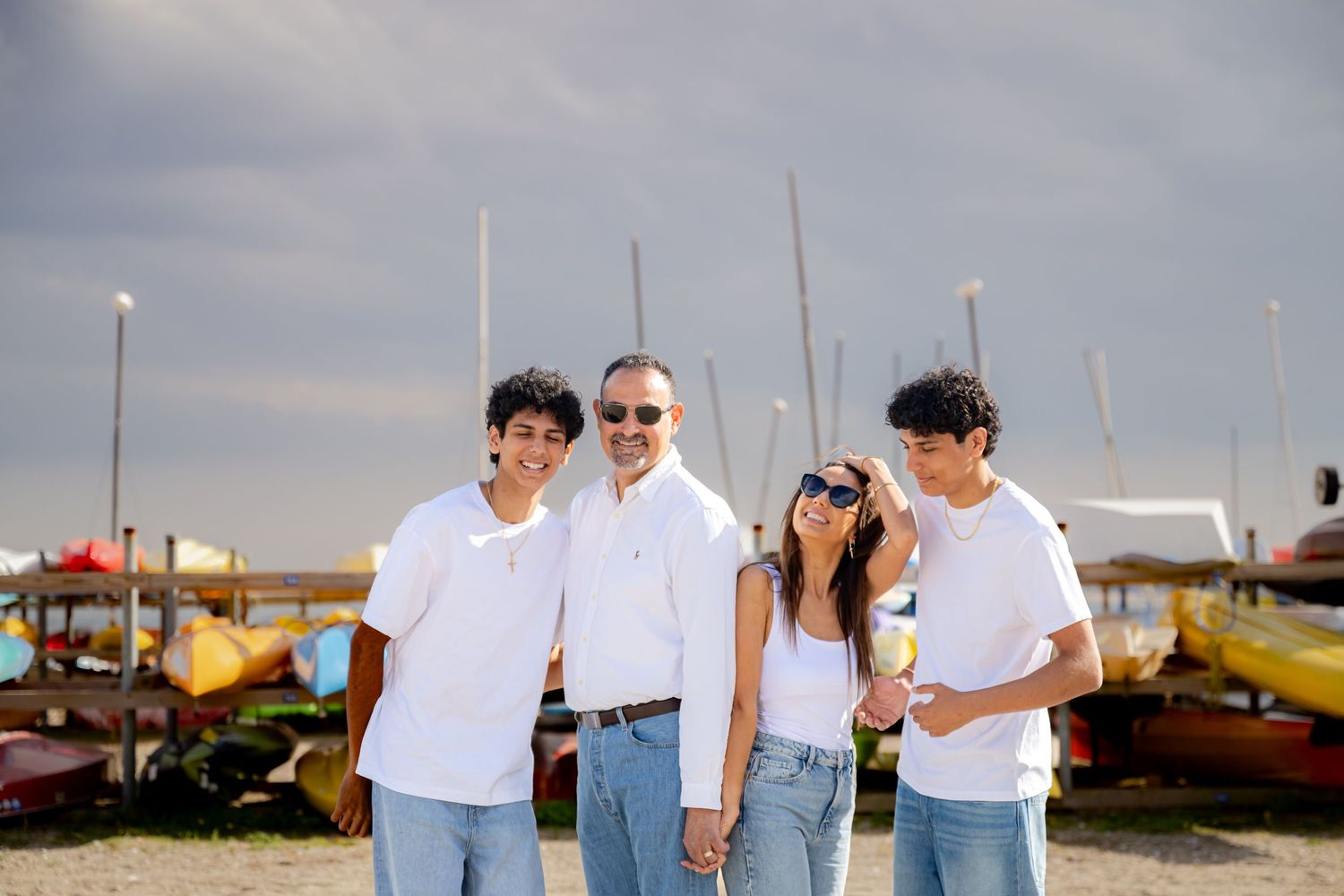 Family of four in white shirts and jeans posing at a marina with colorful boats under cloudy skies.