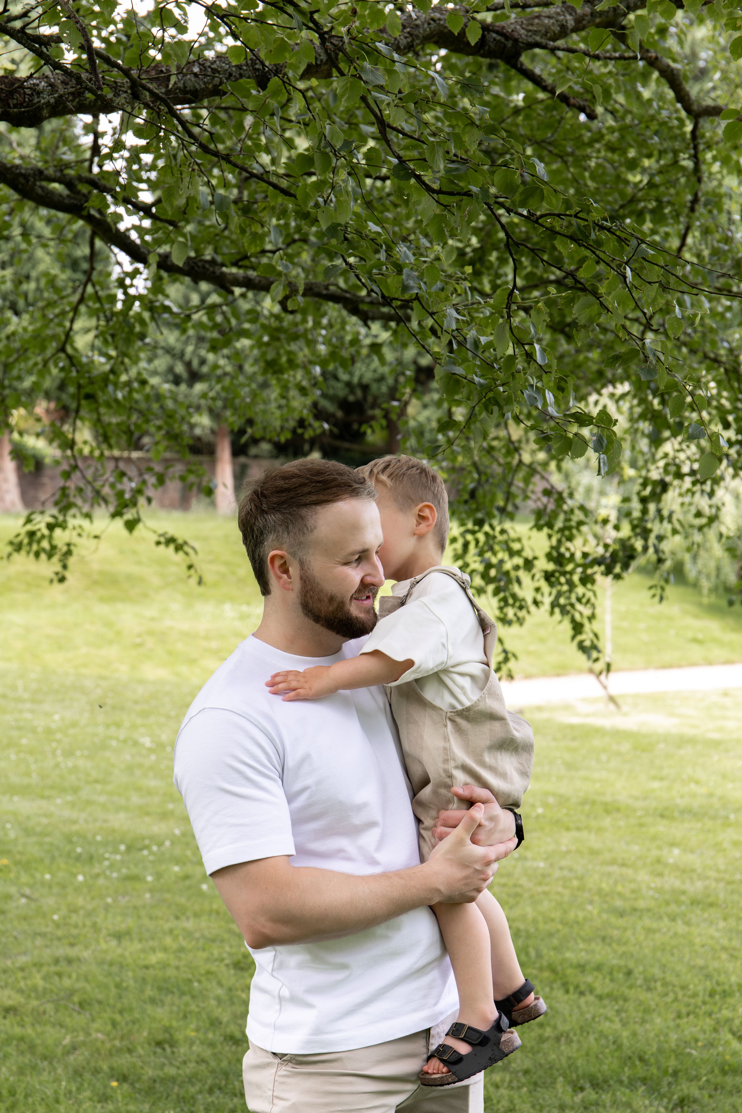 A father holds his young son in a lush green park, sharing a tender moment under a tree Outdoor family photoshoot Nottingham