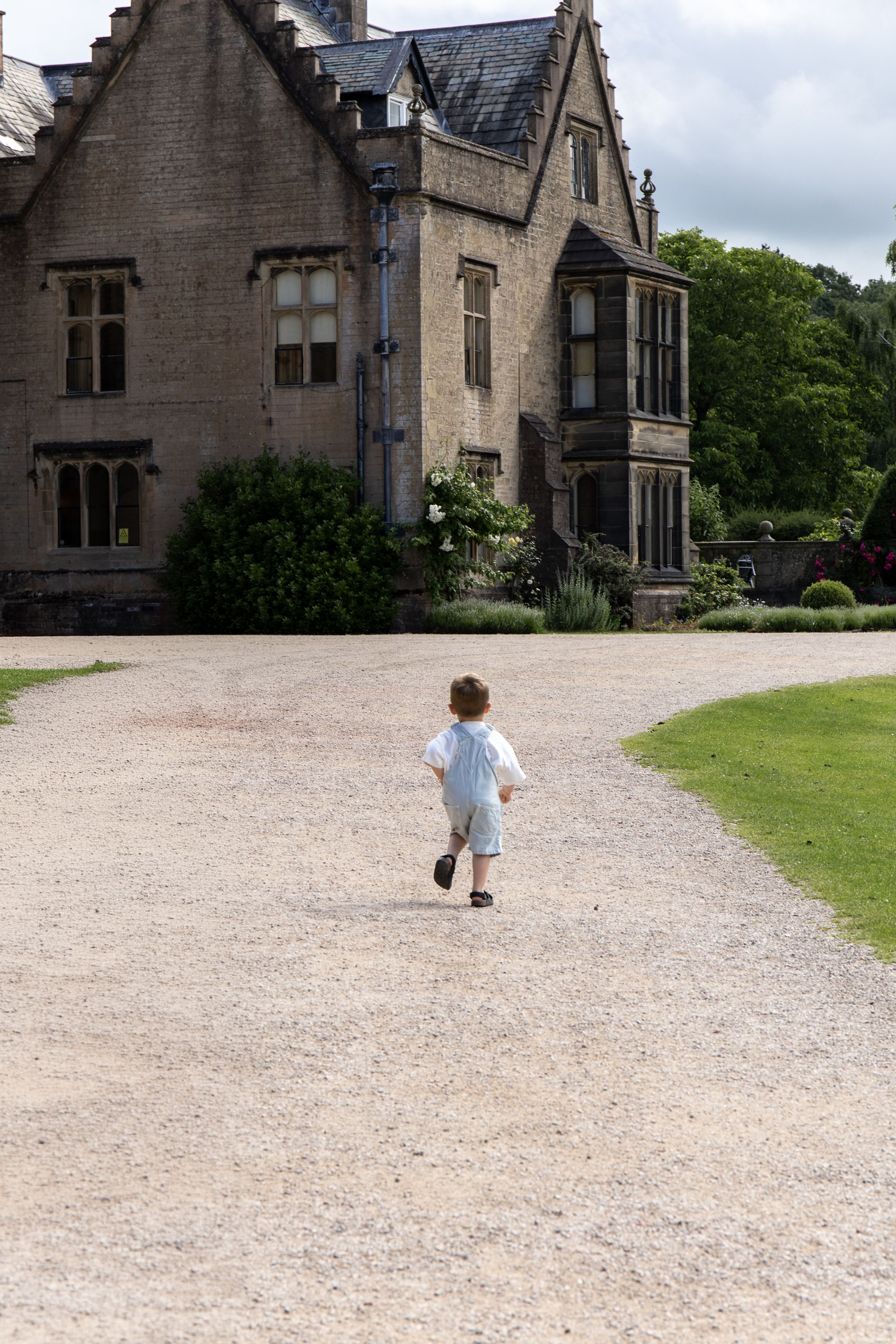 Young child running toward a grand stone manor house along a gravel driveway surrounded by lush greenery Outdoor family photoshoot Nottingha