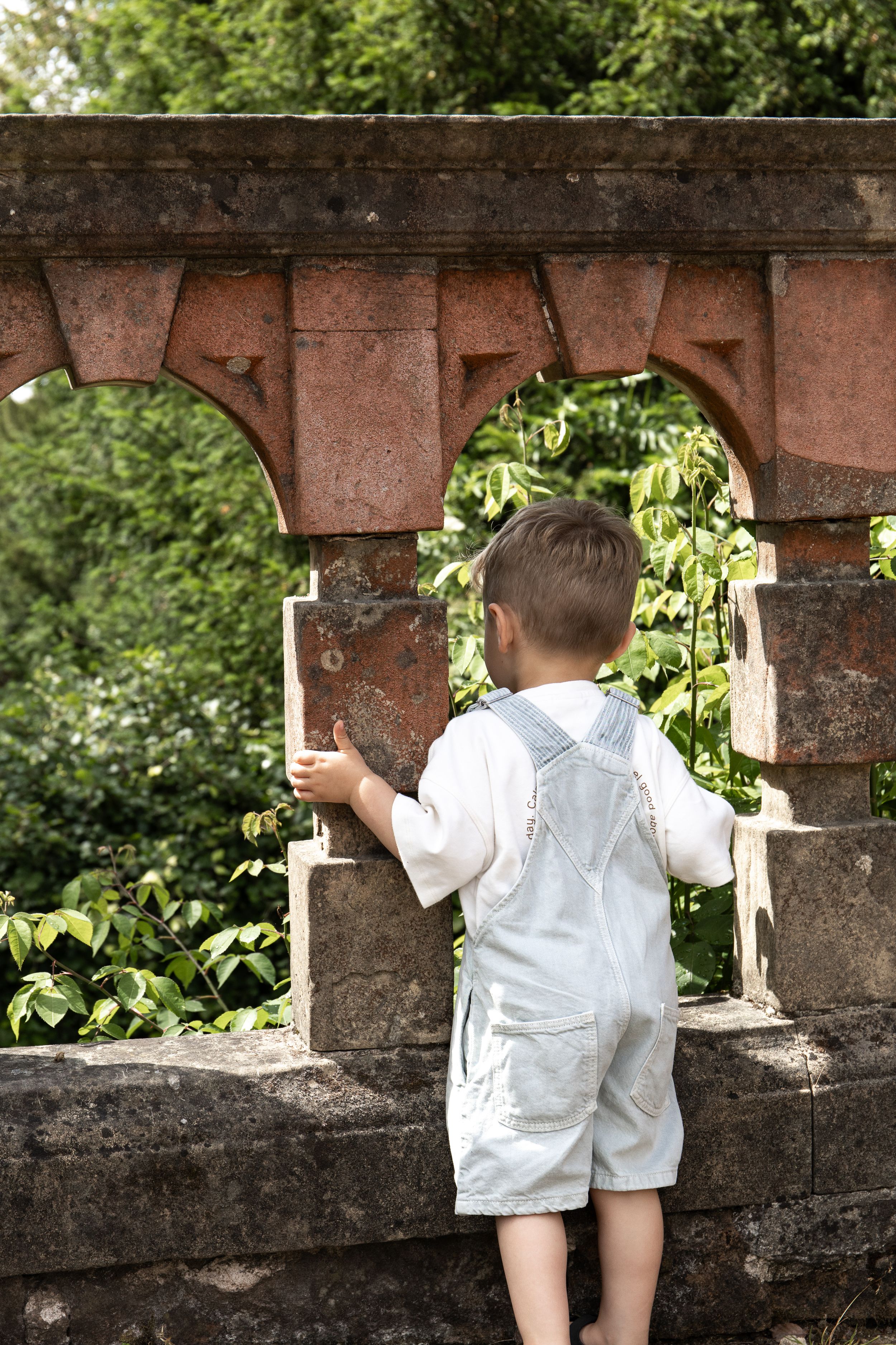 A young boy in white dungarees stands at a stone arch wall, surrounded by lush greenery Outdoor family photoshoot Nottingham