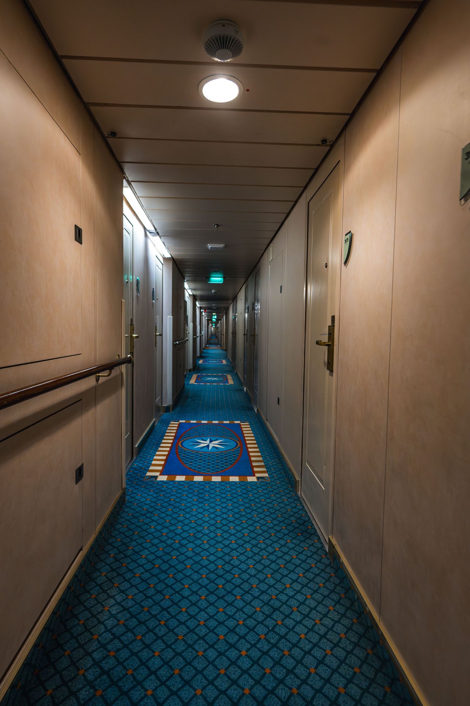 Long cruise ship hallway with blue patterned carpet, cream walls, and doors stretching into the distance.