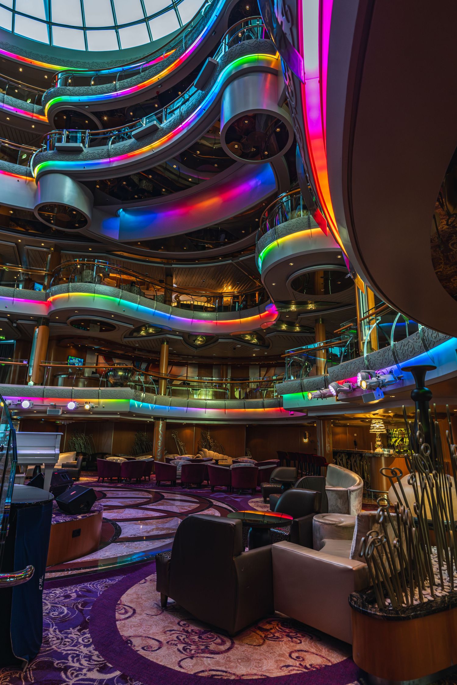 Colorful LED-lit multi-deck atrium lounge on a luxury cruise ship with vibrant rainbow balconies and plush seating.