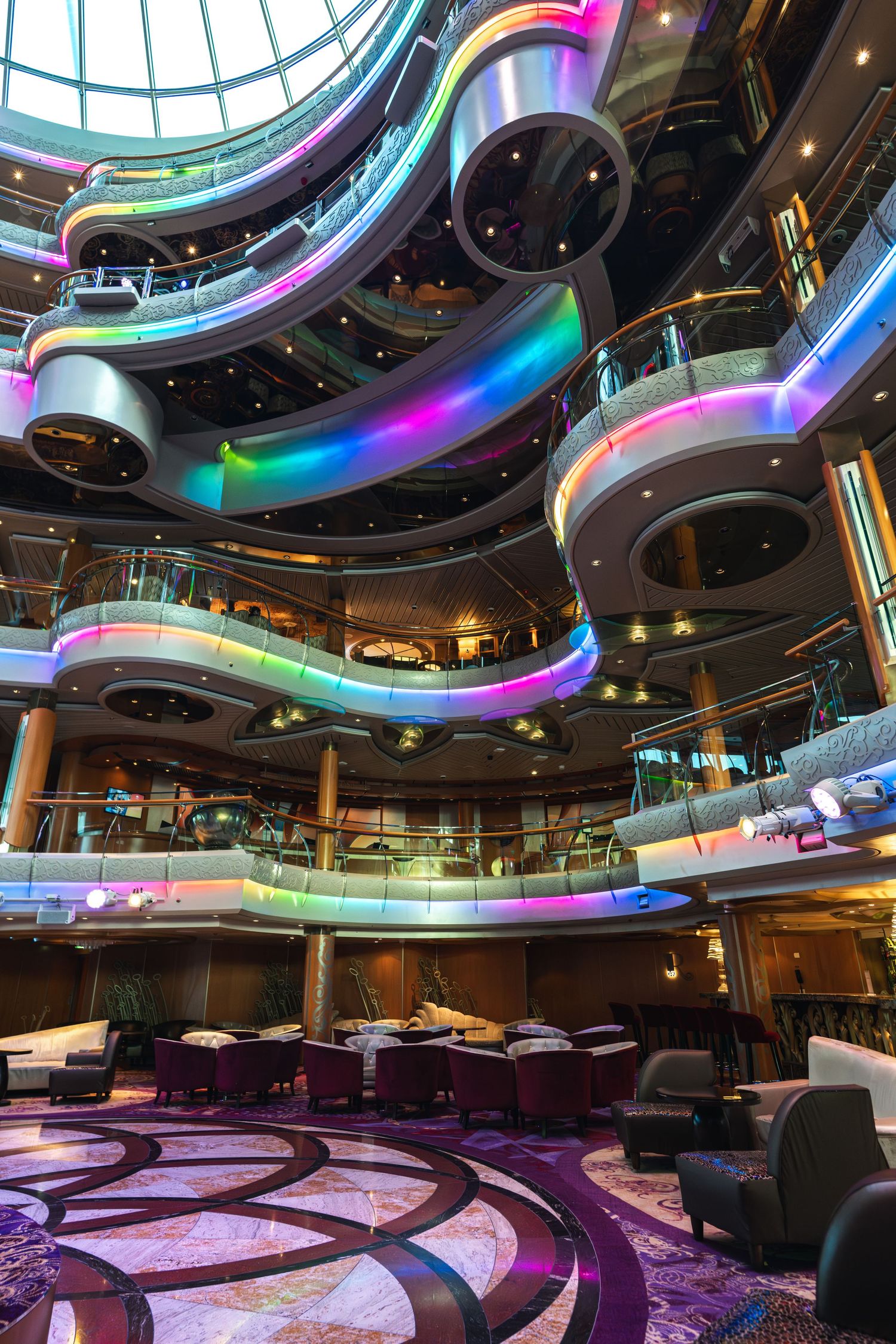 Colorful LED-lit multi-level atrium of a luxury cruise ship with curved balconies and an elegant dining area below.