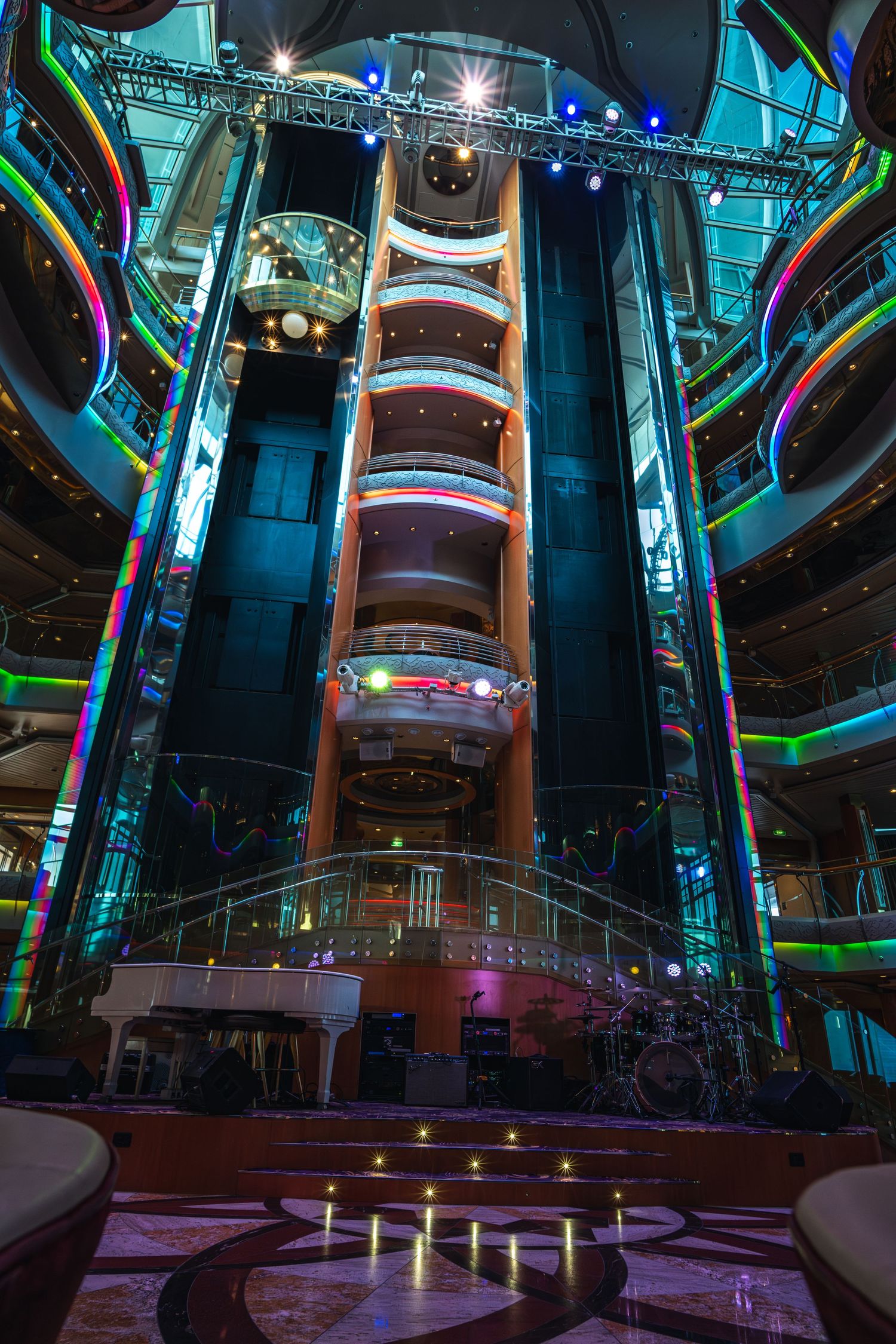 Colorful illuminated atrium on a cruise ship featuring glass elevators, multi-level balconies, and a stage with drum kit.