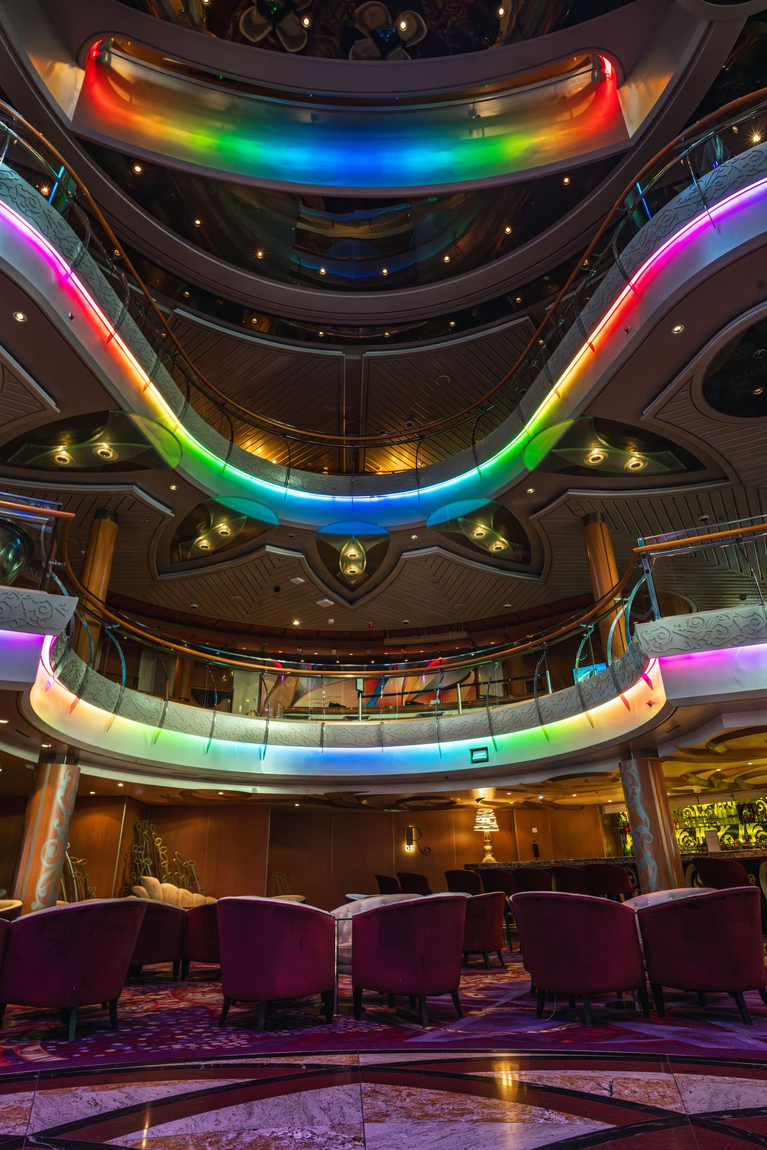 Colorful rainbow LED lights illuminate the multi-level atrium of a luxury cruise ship interior.
