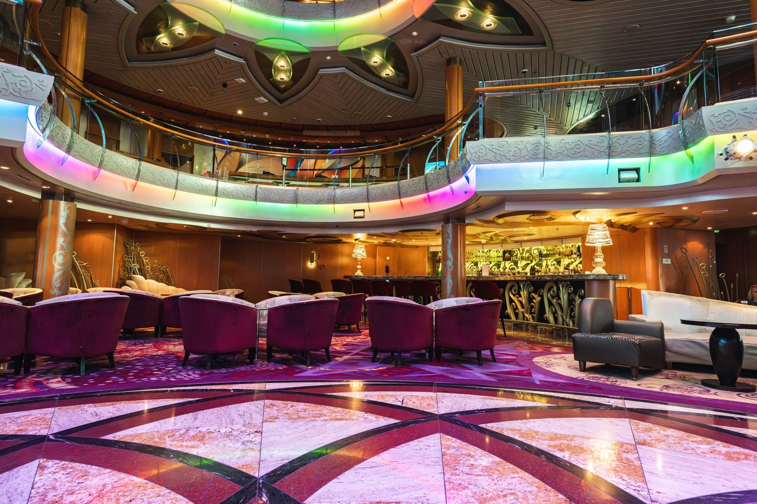 Elegant cruise ship lounge with purple chairs, ornate flooring, multi-level balconies, and colorful ambient lighting.