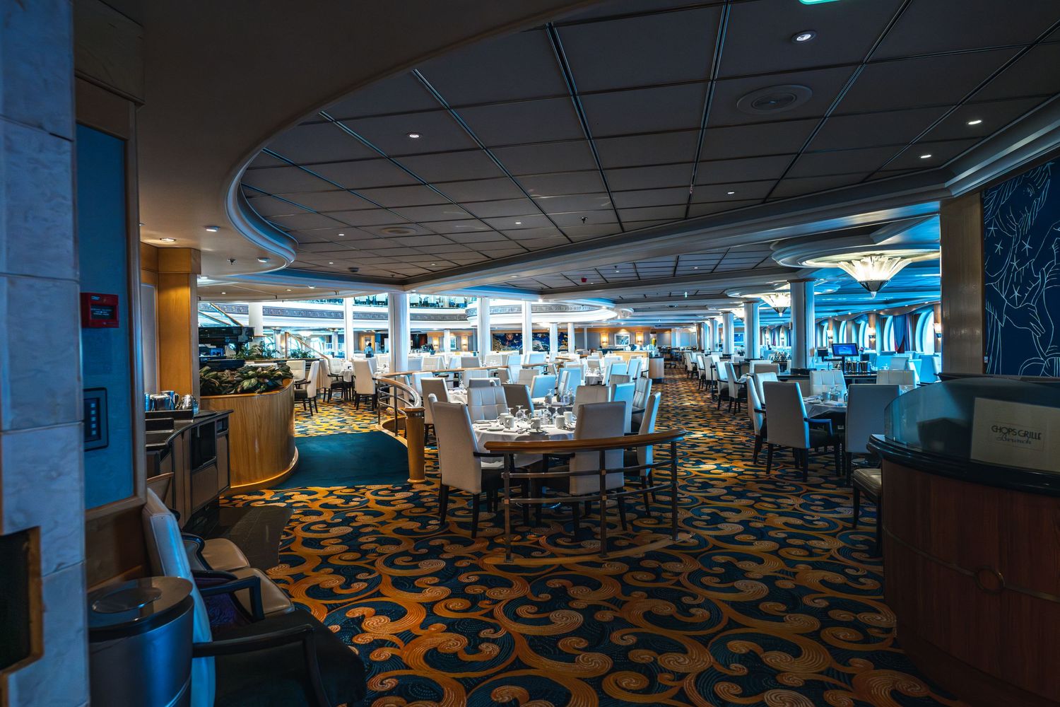 Elegant cruise ship dining room with white chairs, set tables, blue accents, and large windows overlooking the ocean.