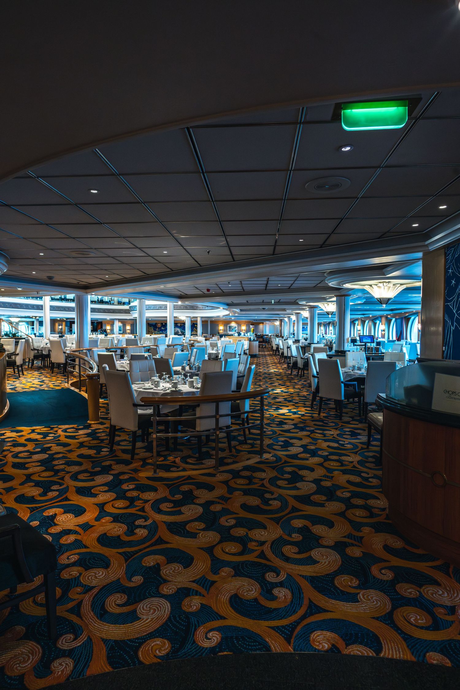 Spacious cruise ship dining room with elegant tables, chairs, and ornate blue and gold patterned carpet.