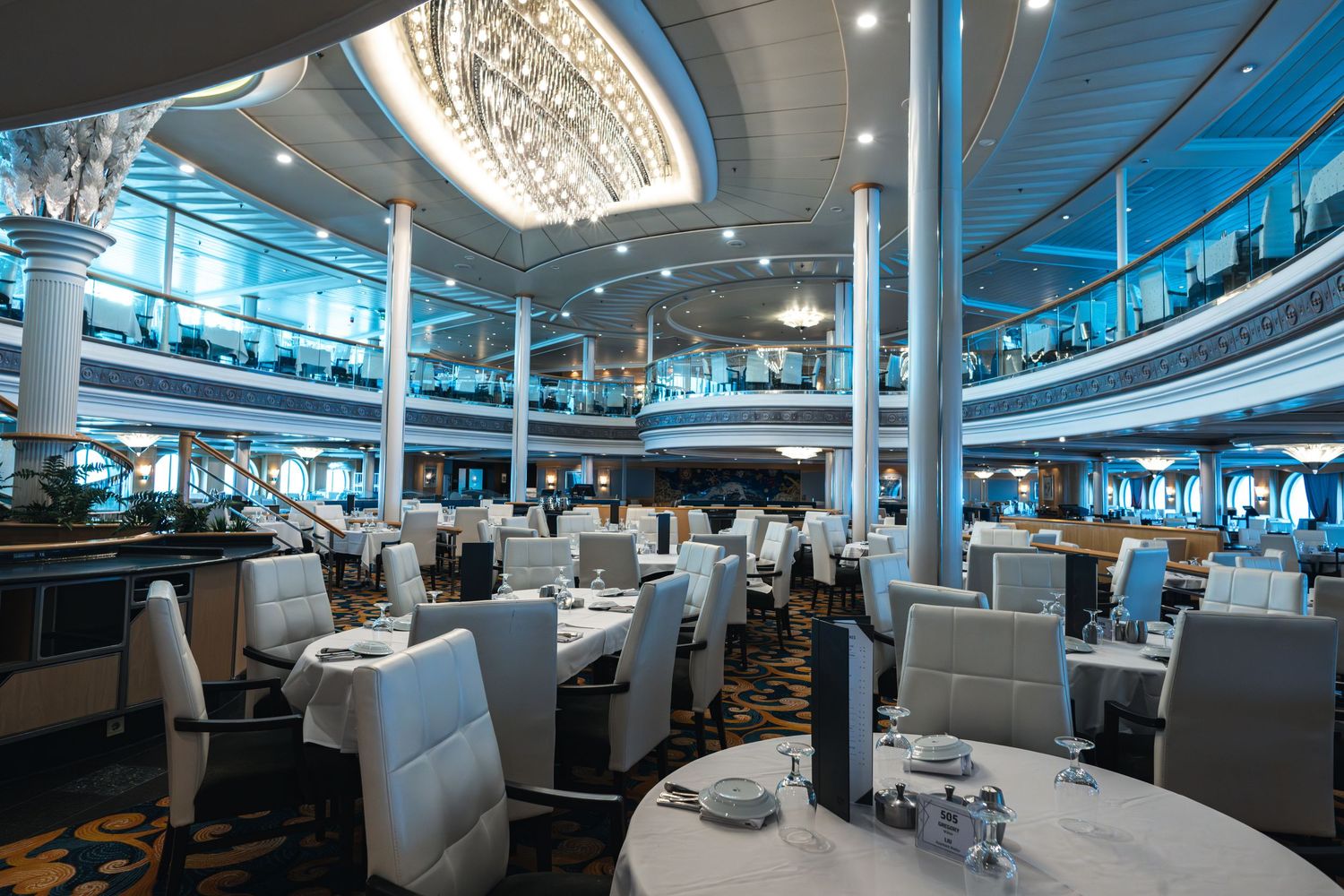 Elegant cruise ship dining room with white chairs, set tables, and a grand chandelier under blue ambient lighting.