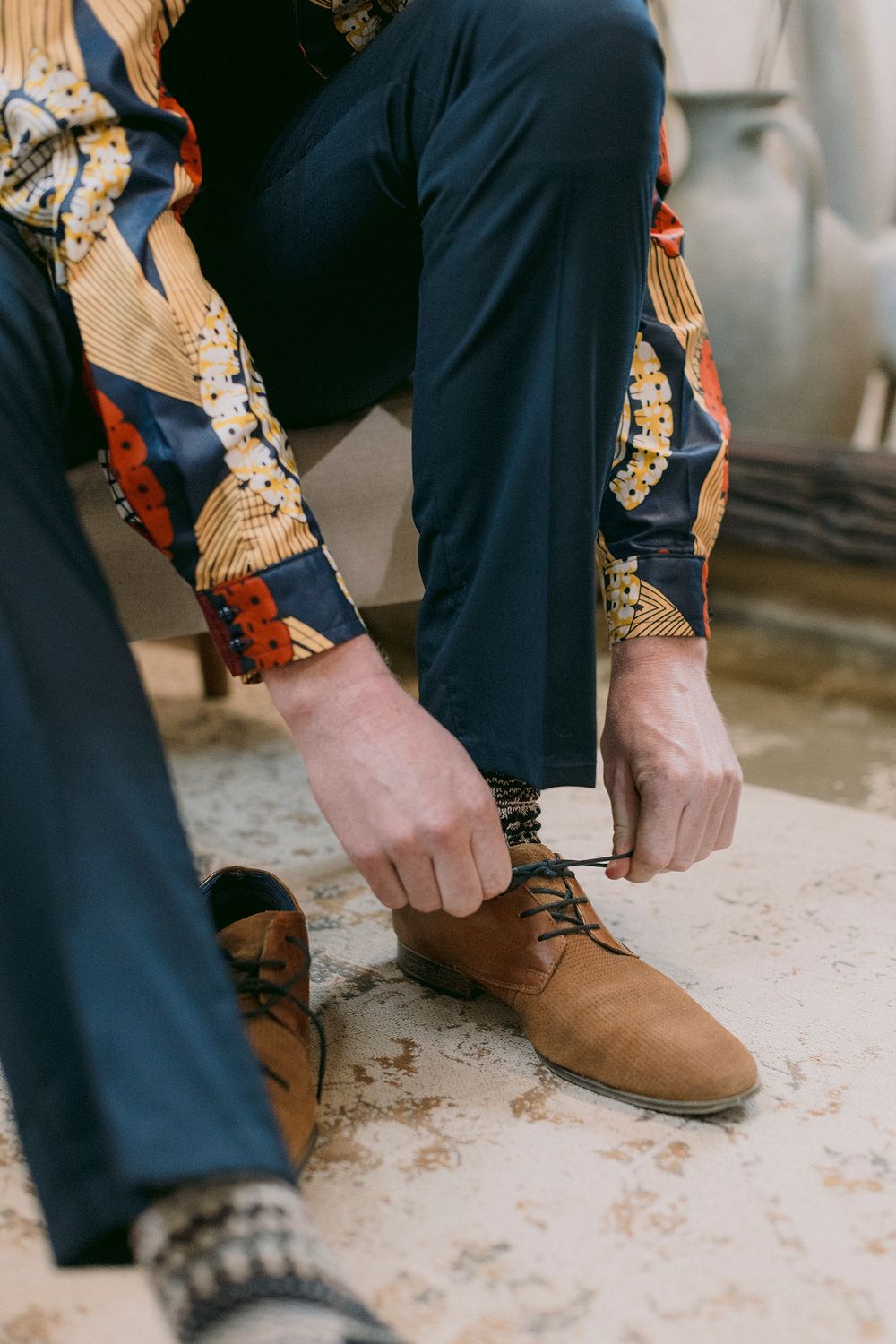 Man in colorful patterned shirt and navy trousers tying laces on brown suede Oxford shoes.