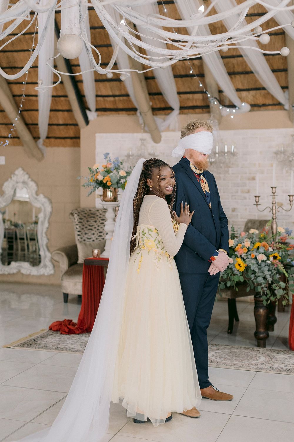 Interracial couple at wedding venue, bride in cream tulle gown leading blindfolded groom in navy suit.