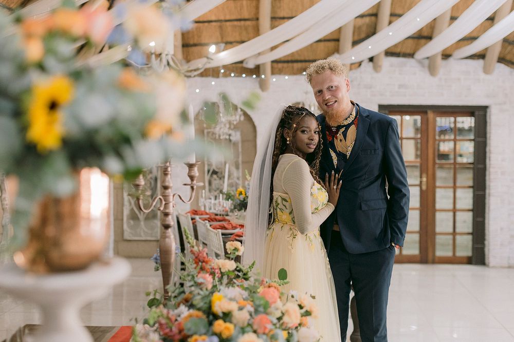 Interracial couple smiling at wedding reception with colorful floral arrangements and sunflowers in elegant venue.